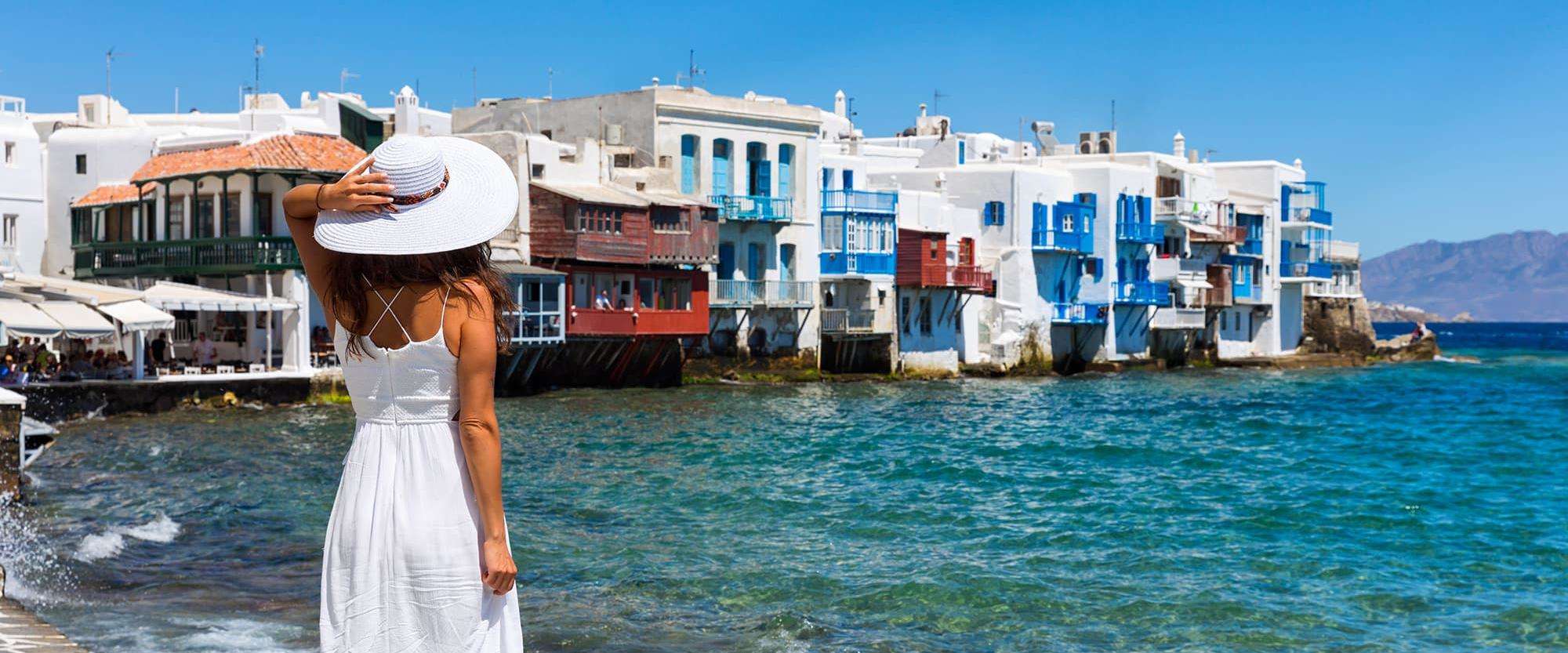 Femme portant un grand chapeau blanc regardant Mykonos et ses bâtisses bleues et blanches au-delà d’une mer bleue